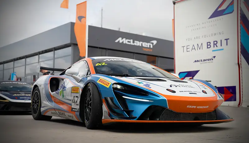 McLaren Artura GT4 racing car. front on photo shot low down the car is orange blue and white. There is a black mclaren building in the background and orange flags. In terms of immersion, these Artura Pro wheels truly evoke the feel of being in a real McLaren GT4 car